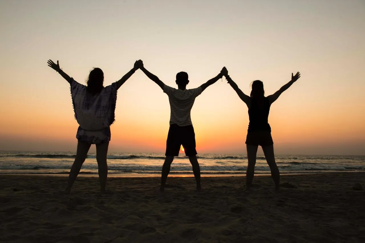 Three people silhouetted against a sunset on a beach, with arms raised.