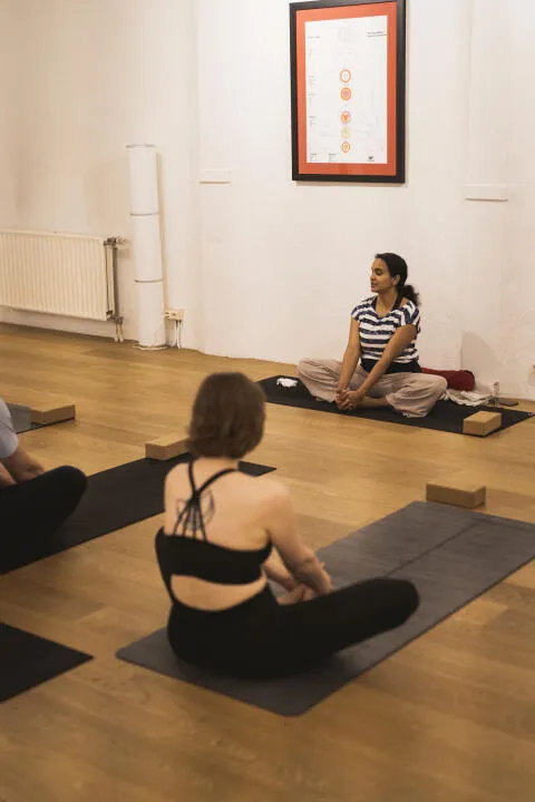 Yoga teacher instructing a class in bound angle pose (baddha konasana) in front of a chakra chart.