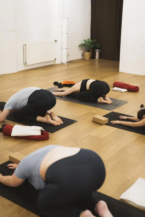 Yoga students resting in child's pose (balasana) on mats in a bright studio.