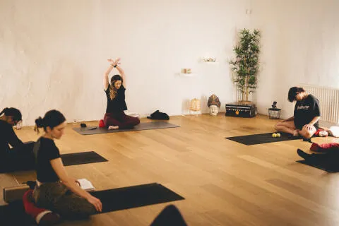 Yoga teacher and students seated on mats practicing an overhead arm stretch during a studio warmup session.