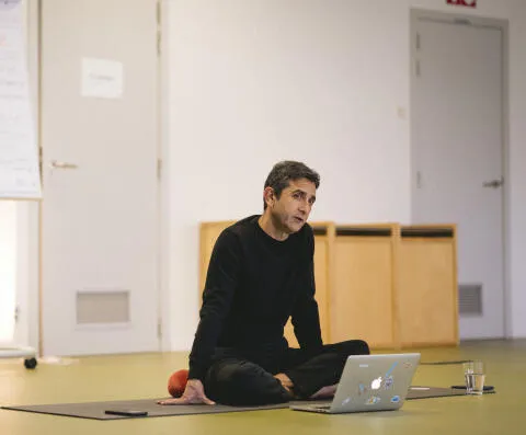 A male yoga teacher sitting cross-legged on a mat using a laptop during a studio lecture.