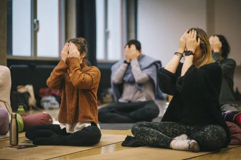 People sitting cross-legged in a studio covering their eyes with their hands during a yoga or meditation class.