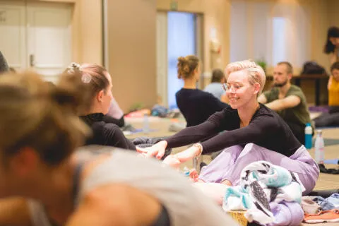 A smiling woman with short blonde hair practicing partner yoga stretches in a studio class.