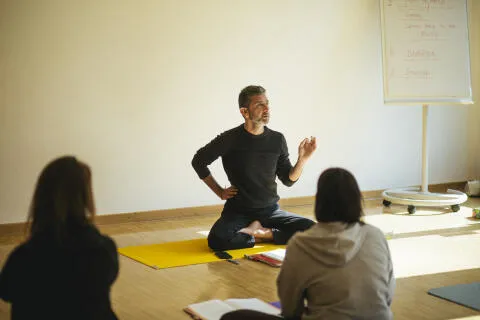 A male instructor sitting cross-legged on a yellow mat teaching yoga philosophy to students.